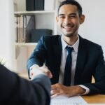 Confident businessman in suit shaking hands at office desk, symbolizing successful partnership.